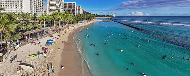 This aerial shot captures the vibrant energy of Waikiki Beach, Hawaii, where turquoise waters meet golden sands. Tourists bask in the sun, swim, and enjoy the lively atmosphere, framed by swaying palm trees and the iconic skyline of Honolulu.