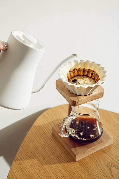 Hot water from a kettle is being poured into V60 pour-over funnel that sits atop carafe, allowing alternative method of coffee brew.