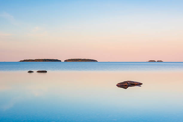 At Lake Vanern in Sweden, the morning light casts a gentle glow on the still water, creating a serene atmosphere. Rock formations emerge from the lake, while distant islands fade into the horizon.