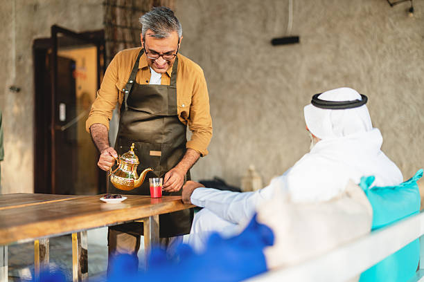 A middle-aged Middle Eastern waiter in a tan shirt and apron serves tea from a golden teapot to a customer in traditional attire at a cozy Al Fahidi cafe. Sunlit and warm ambiance enhances the scene.