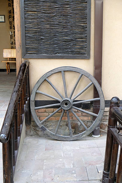 An antique wooden wheel leans against a textured wall next to a staircase in a rustic interior. The warm tones create a nostalgic atmosphere