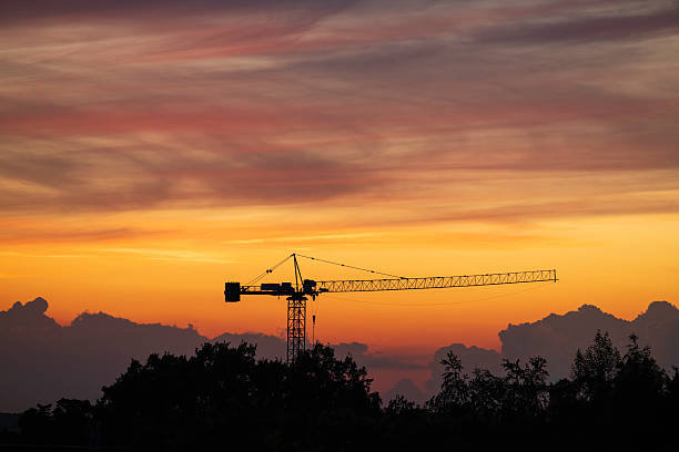 A construction crane stands tall against a vibrant sunset sky filled with clouds. The scene captures the beauty of nature amidst urban development during twilight.