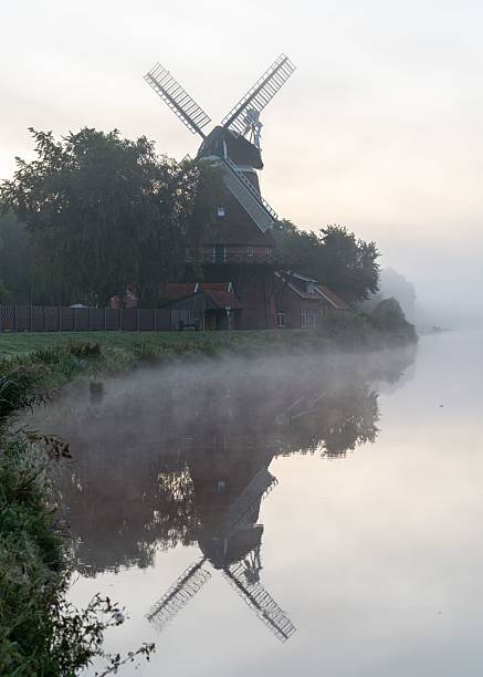 Scenic view of a traditional windmill reflecting in a misty river at dawn, surrounded by lush greenery, creating a serene and tranquil atmosphere.
