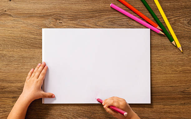 Top view of child a drawing with a pencil on blank paper, colorful crayons nearby on wooden table