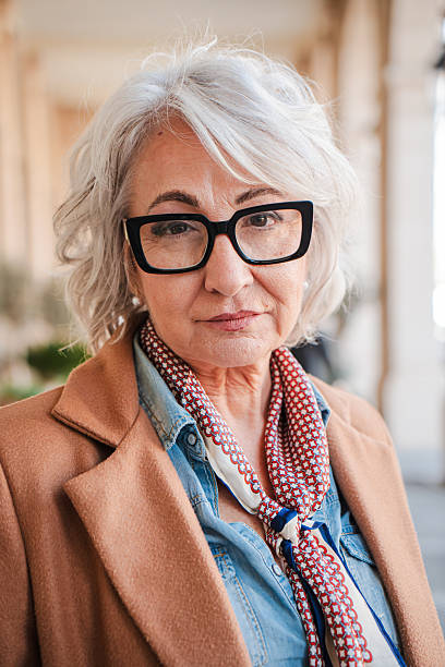 Confident elderly woman with silver hair and glasses looking directly at the camera while expressing a serious demeanor in a thoughtfully composed portrait photograph