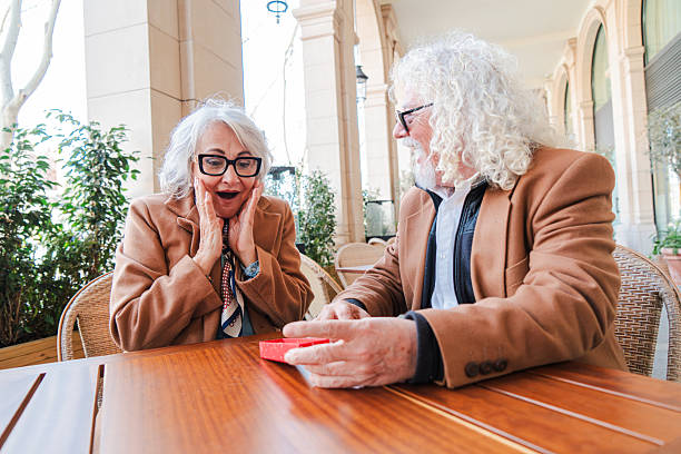 Joyful seniors sharing an exciting moment while exchanging a delightful surprise gift at a romantic terrace table during a charming date full of laughter and love, celebrating their special bond together