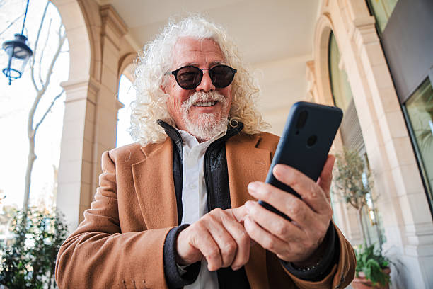 istock Cheerful older man with curly hair happily using his smartphone while smiling and engaging with a mobile application outdoors, showcasing the joy of technology and connectivity in everyday life 2207699109