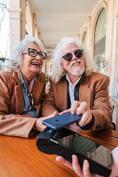istock Joyful senior couple happily using smartphone to make a payment while enjoying their time together at a cozy cafe table with bright smiles and engaging expressions on their faces and serenity in the air 2207699472