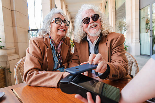 Joyful elderly couple happily engaging in mobile payment transaction using smartphone and credit card at a cozy cafe table while sharing smiles and laughter together in delightful companionship