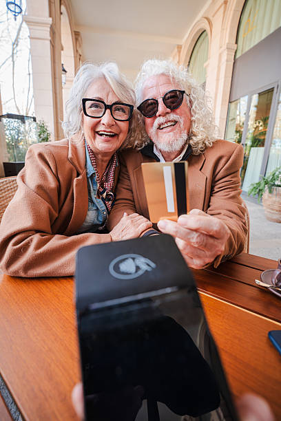 istock Cheerful elderly couple happily interacting with their credit card while enjoying a delightful meal together at a charming cafe, showcasing their joyful spirit and connection over a financial transaction 2207700486