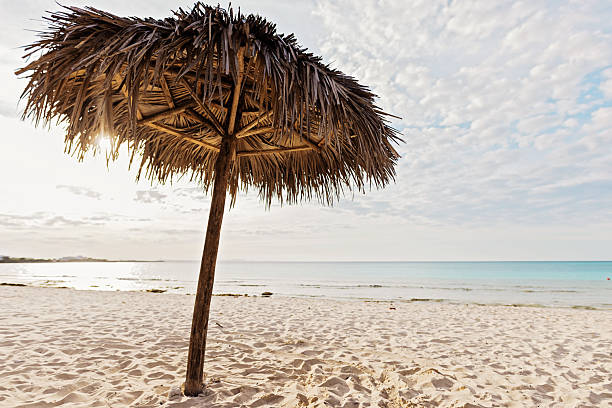 A picturesque tropical beach with a straw parasol, white sand, and a tranquil sea under a sunny, partly cloudy sky.