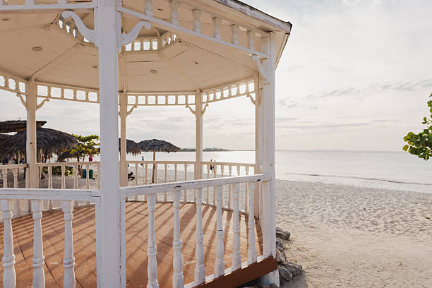 Serene view of a white wooden gazebo on a sandy beach, surrounded by a calming ocean and sunset. A peaceful tropical scene ideal for vacation, relaxation, or travel themes.