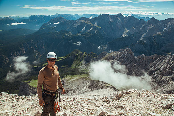 A climber in protective gear stands triumphantly at a high altitude overlooking the breathtaking mountain scenery of Austria. Clouds weave through the valleys, enhancing the view.