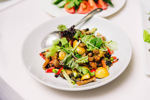 Colorful mixed salad with grilled vegetables, tender meat pieces, potatoes, and leafy greens served on a white plate with spoon.