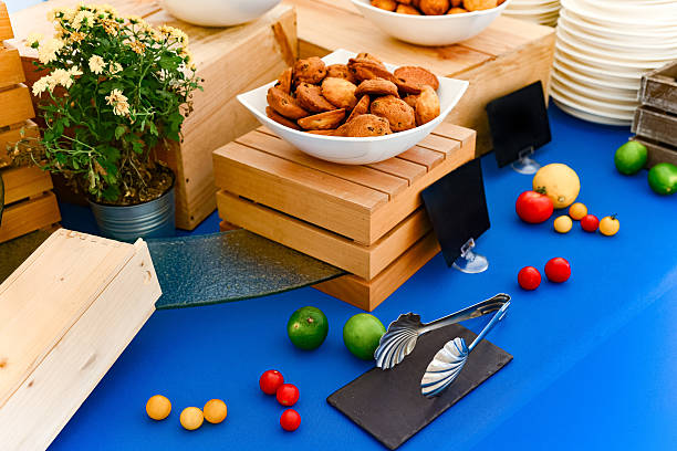A white bowl filled with cookies is displayed on a wooden riser, surrounded by fresh fruits and floral decor at an event.