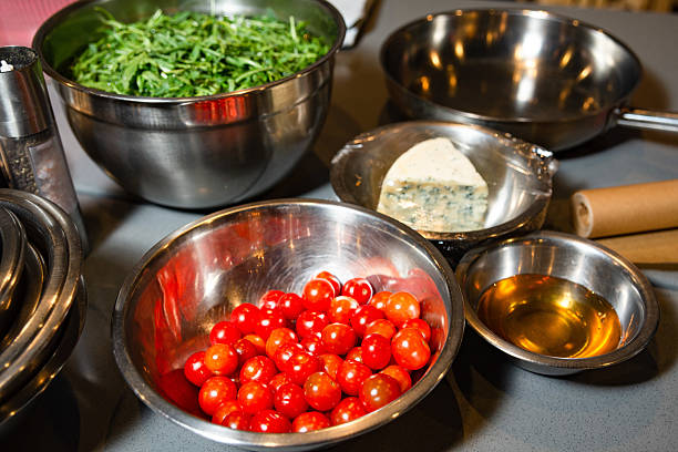 Metal bowls with fresh cherry tomatoes, arugula leaves, blue cheese, and dressing ready for preparing a healthy salad dish.