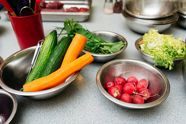 Assorted fresh vegetables including carrots, cucumbers, radishes, and greens arranged in metal bowls on a kitchen counter for cooking.