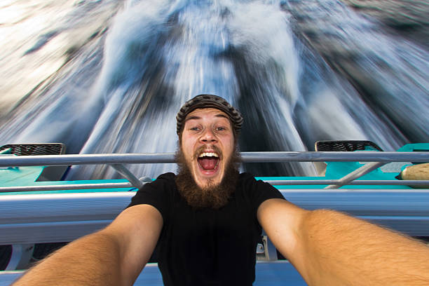 A man with a beard is smiling widely while holding a selfie stick on a speedboat, surrounded by water splashing in the background. It's a bright, sunny day.