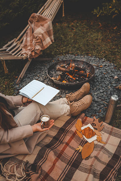 istock Woman writing in a notebook by the campfire, holding a cup with marshmallows. Cozy autumn setup with blanket, croissant, thermos, and fall leaves 2207732164