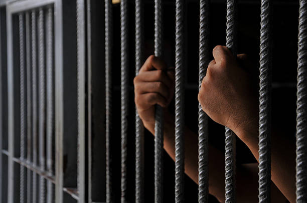 istock prisoner's hands behind bars with black background 2207734293