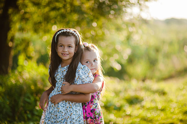 Two young Asian girls bonding their siblings relationship while embracing each other as they have fun and enjoy the good weather together in the country park, . Two female children hugging, talking and laughing while resting and sitting on the ground of leaves in the woods, under the sun with trees during Autumn.