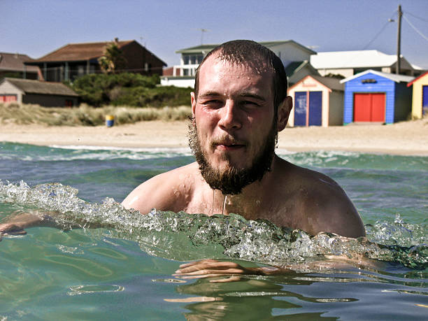A young man with a beard is swimming in the clear blue waters of an Australian beach. Colorful beach huts line the shore under a bright sky, creating a cheerful atmosphere.
