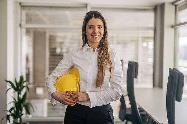 istock Female engineer smiling and holding yellow hardhat in office 2207744840