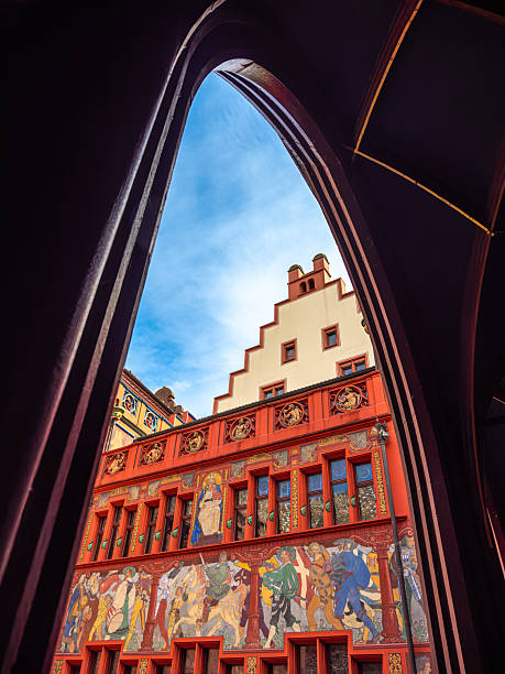 Basel, Switzerland - August 17, 2024: A vibrant architectural scene featuring a historic city hall of Basel with ornate frescoes and a stepped gable, viewed through an archway against a blue sky.