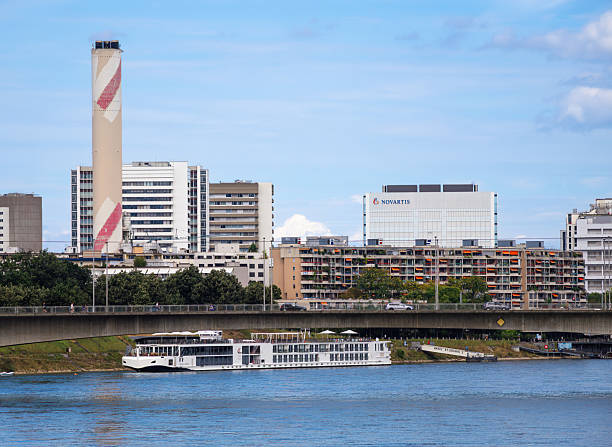 Basel, Switzerland - August 17, 2024: A river cruise ship sails under a bridge in an urban area with modern buildings, including a tall chimney and a building with the Novartis logo.