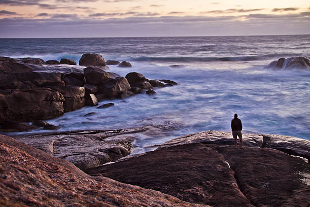 A person stands on large rocks at the shore, gazing at the waves during sunset in Western Australia. The ocean is calm, with gentle waves lapping against the rocks as the sky fades into dusk.