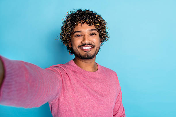 istock Happy young man with curly hair wearing pink pullover taking a selfie against a blue background 2207750848