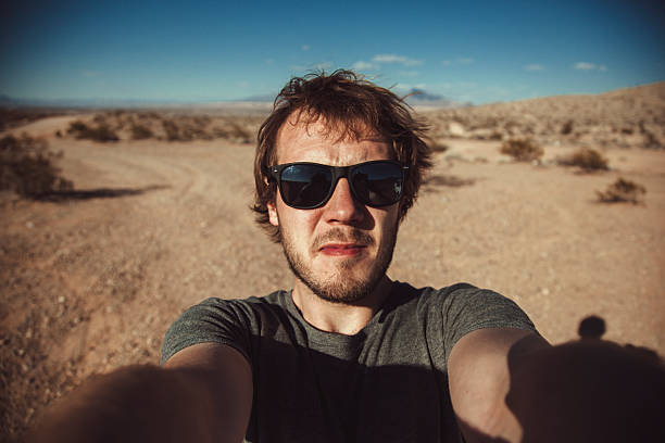 A man stands in the Nevada desert, wearing sunglasses and a gray shirt, taking a selfie with a scenic background of arid land and clear blue skies, enjoying the sunny weather.