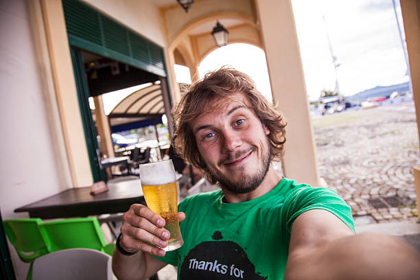 A cheerful man poses for a selfie while holding a cold beverage outside a cafe. The picturesque waterfront setting features boats and a vibrant atmosphere under a blue sky.