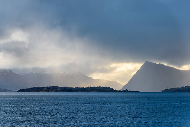 Dramatic clouds gather over serene mountain landscape by the sea during early evening hours