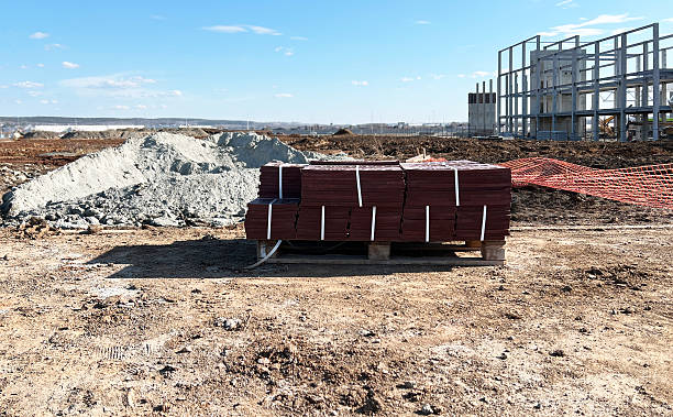 istock Construction site showcasing neatly stacked red tiles on pallets and a partially built structure in the background. Sandy ground and protective netting visible, indicating active work 2207767049