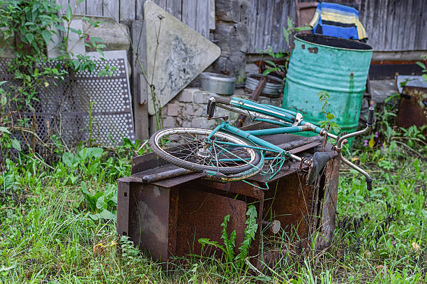 A vintage bicycle lies abandoned and rusting in an overgrown backyard. Surrounded by greenery and discarded items, the scene evokes a sense of nostalgia and neglect.