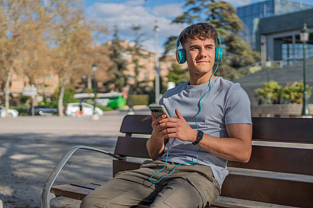 Young man sits on a park bench, enjoying music through his headphones and holding his smartphone, taking a break in the urban environment