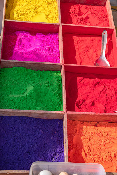 Bright cups of colored Holi powders arranged in a red basket on a street, with a person seated nearby. A vivid display of pigment, color, and Indian festival atmosphere.