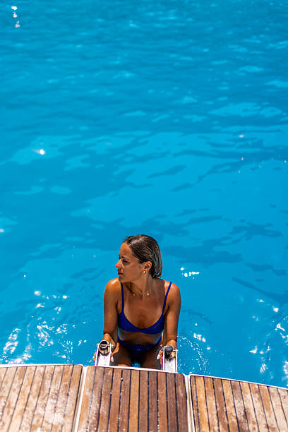 Attractive blond woman in a blue bikini is climbing ladders on a sailboat, getting out of the sea. The sea is beautiful turquoise color.