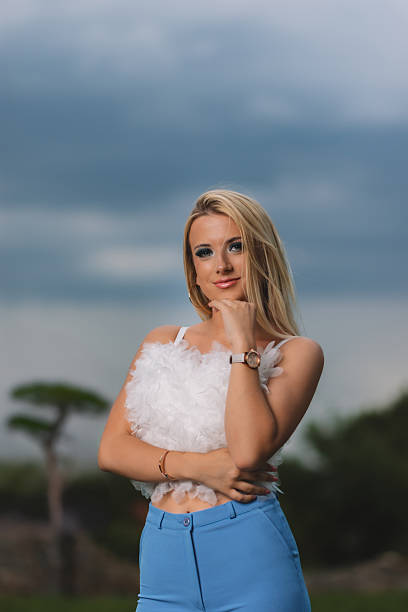 A fashionable young woman with blonde hair posing confidently in an outdoor setting. She wears a trendy white feathered crop top and high-waisted blue pants, accessorized with a watch and bracelet. The blurred green background and natural lighting create a vibrant and professional portrait, perfect for fashion, lifestyle, or influencer-related content.
