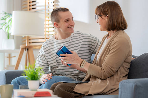 A cheerful mother sits on a comfortable sofa, smiling warmly as she opens a present from her child on Mother’s Day. This touching moment captures family bonding, appreciation, and the heartfelt joy of giving and receiving gifts in a cozy home setting.