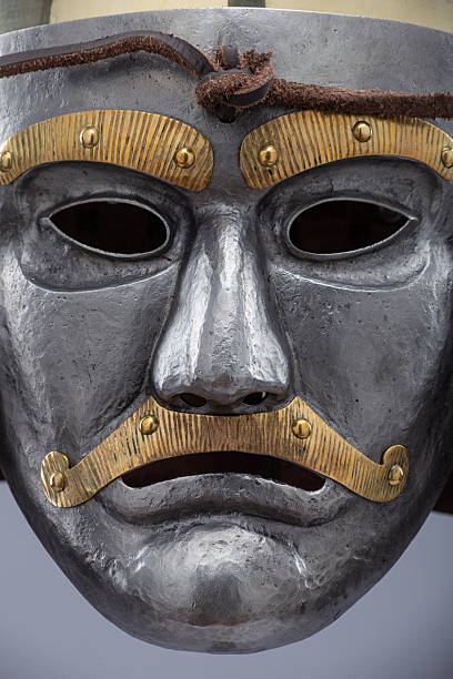 istock Historical metallic combat helmet with integrated face mask. Pointed steel helmet featuring a detailed metallic face mask with eye openings, decorative elements, and side protection flaps. 2207797455