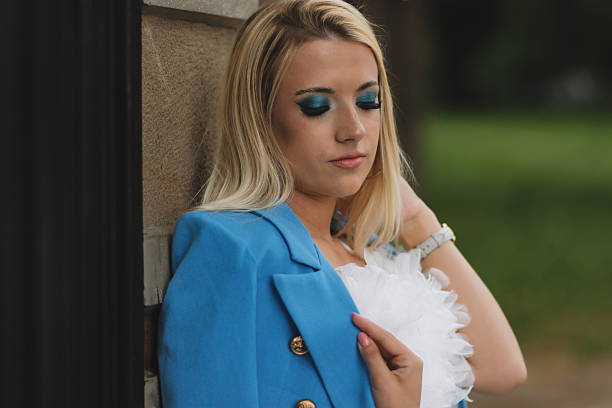 A modern young woman with long blonde hair poses confidently leaning against a stone gate. She is wearing a trendy white feathered crop top and vibrant blue pants that exude elegance and modern style. The background features an old brick and stone building, adding depth to the portrait. Ideal for fashion, confidence and lifestyle themes.