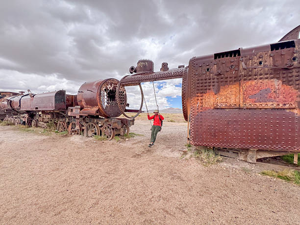 A person in a red jacket swings from an old train boiler in a desert. Unique outdoor play. Adventurous traveler exploring abandoned landscape attractions. Industrial decay in nature.