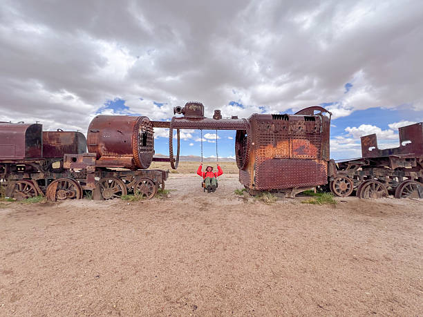 A person in a red jacket swings from an old train boiler in a desert. Unique outdoor play. Adventurous traveler exploring abandoned landscape attractions. Industrial decay in nature.