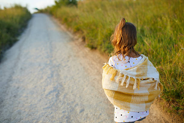 A young girl strolls along a dirt path, enjoying the summer sun. She wears a light shawl over her shoulders and denim shorts, surrounded by green hills and nature.