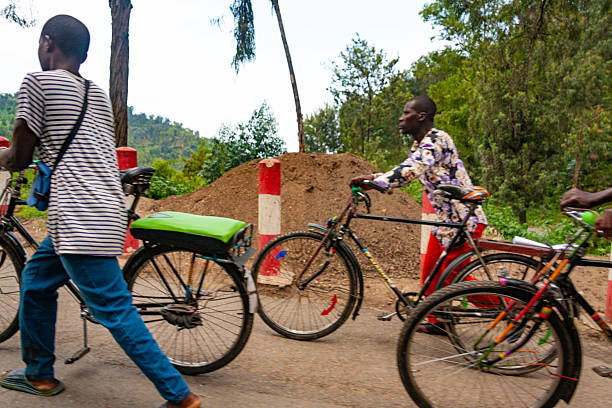 03-27_2025\nIndividuals with bicycles on a road in Rwanda. The image shows several people, including one wearing a striped shirt and blue jeans walking alongside their bicycle, and another wearing a patterned shirt pushing their bicycle. The scene features a pile of earth or sand, and red and white striped poles, suggesting a road construction or maintenance area. The image captures a moment of everyday life and transportation in Rwanda.