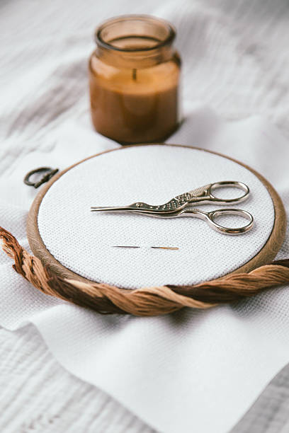 istock Embroidery hoop with white canvas, vintage stork scissors, needles and embroidery thread arranged neatly on soft fabric, candle in background, representing handmade craft, cross stitch, vertical 2207813997
