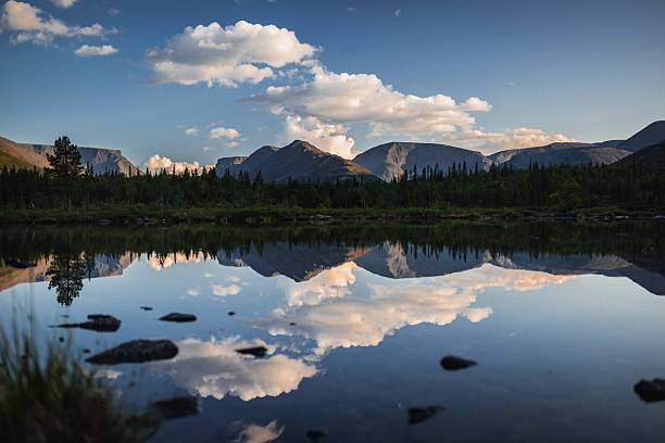 A calm lake reflecting lush forest, majestic mountains, and soft clouds on a sunny day. Nature's untouched beauty is showcased in this peaceful and picturesque landscape, offering a sense of tranquility and awe.