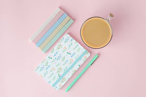 A floral notebook, pastel-colored pens, a green pen, and a cup of coffee on a pink background, symbolizing creativity, organization, and relaxation